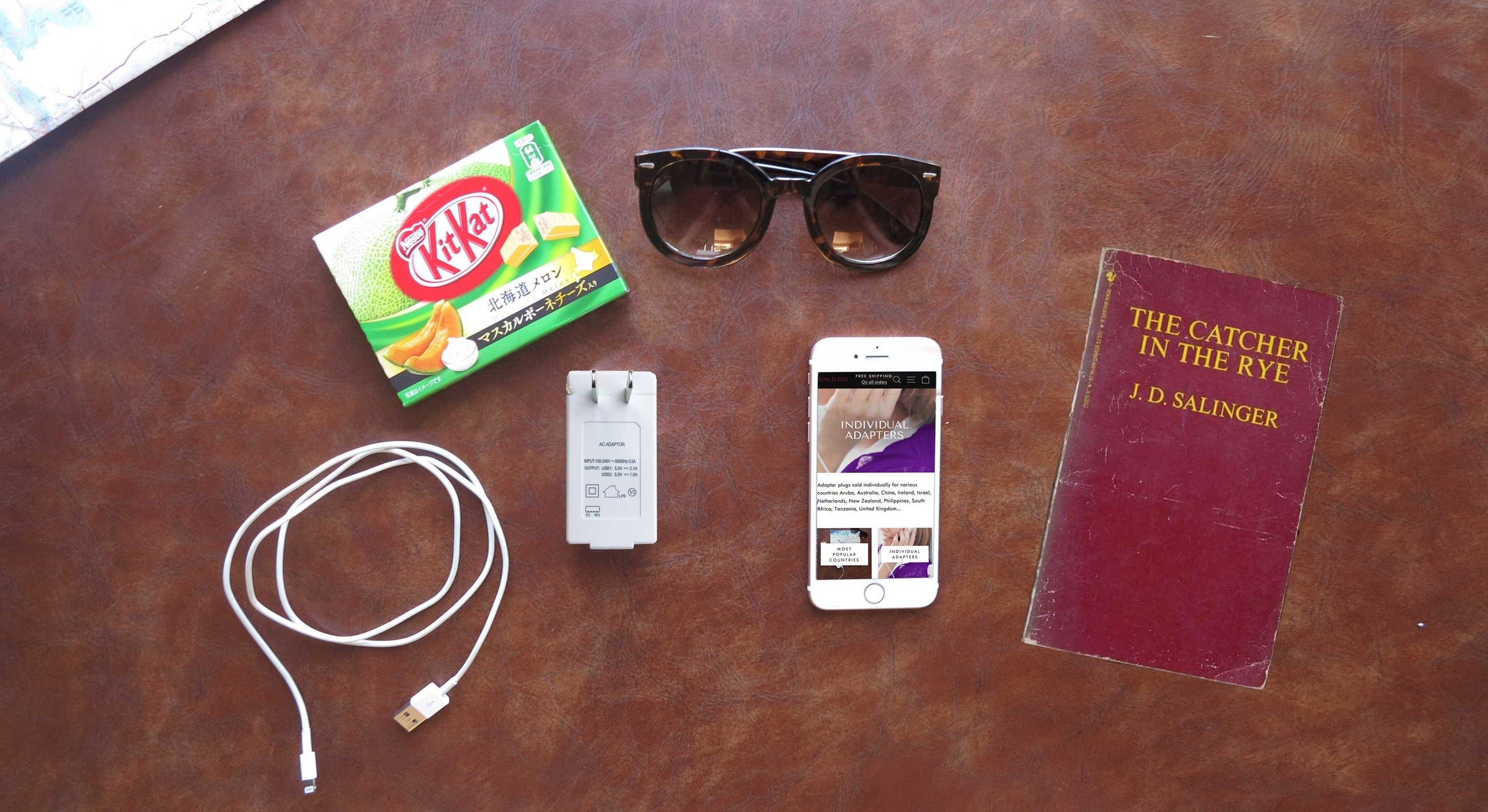 Flatlay of Japan adapter plug iphone, book, sunglasses, charging plug and kit kat