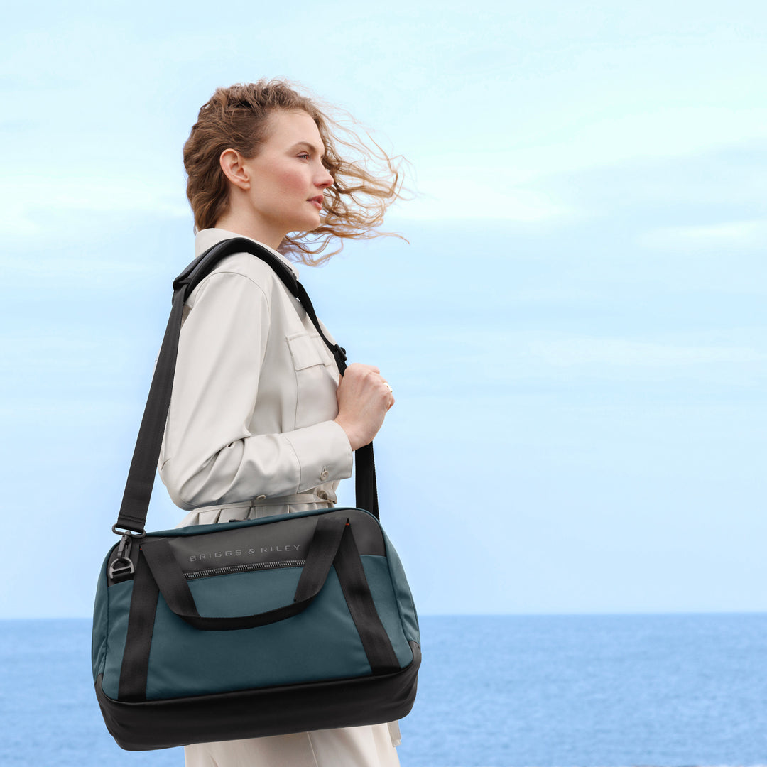 Woman stands on beach with ocean breeze holding Briggs & Riley ZDX Underseat Cabin Bag in color Ocean blue over her shoulder